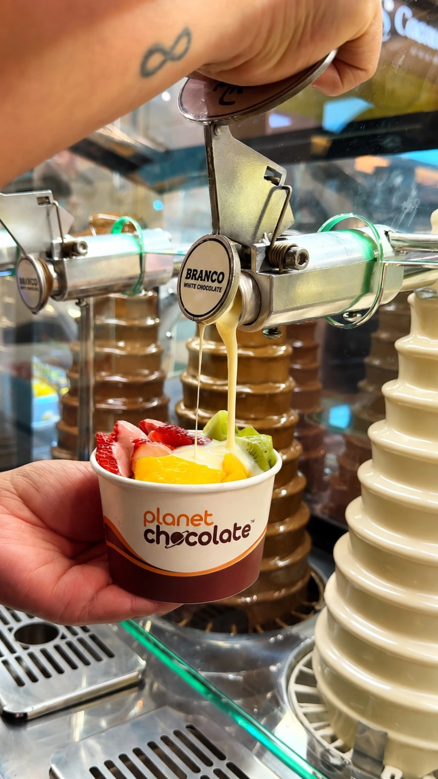 Chocolate branco sendo servido pela cascata sobre frutas em um pote Planet Chocolate.